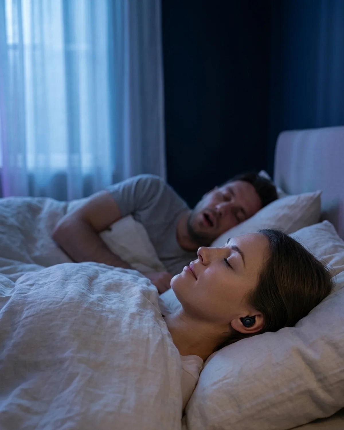 Woman asleep with matte black sleep earplug, partner snoring beside her in dark bedroom